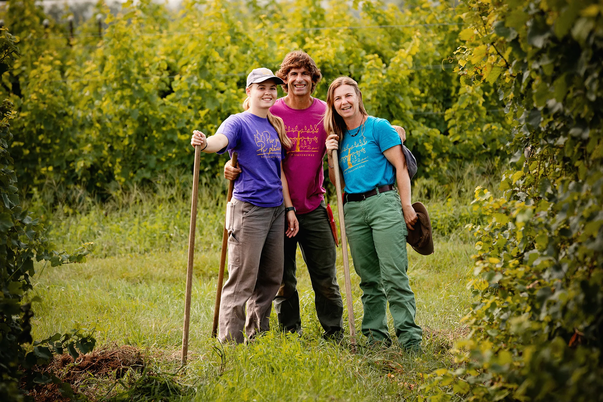 Three smiling people in coloured T-shirts stand arm in arm in a grassy vineyard alley, with long-handled tools beside them