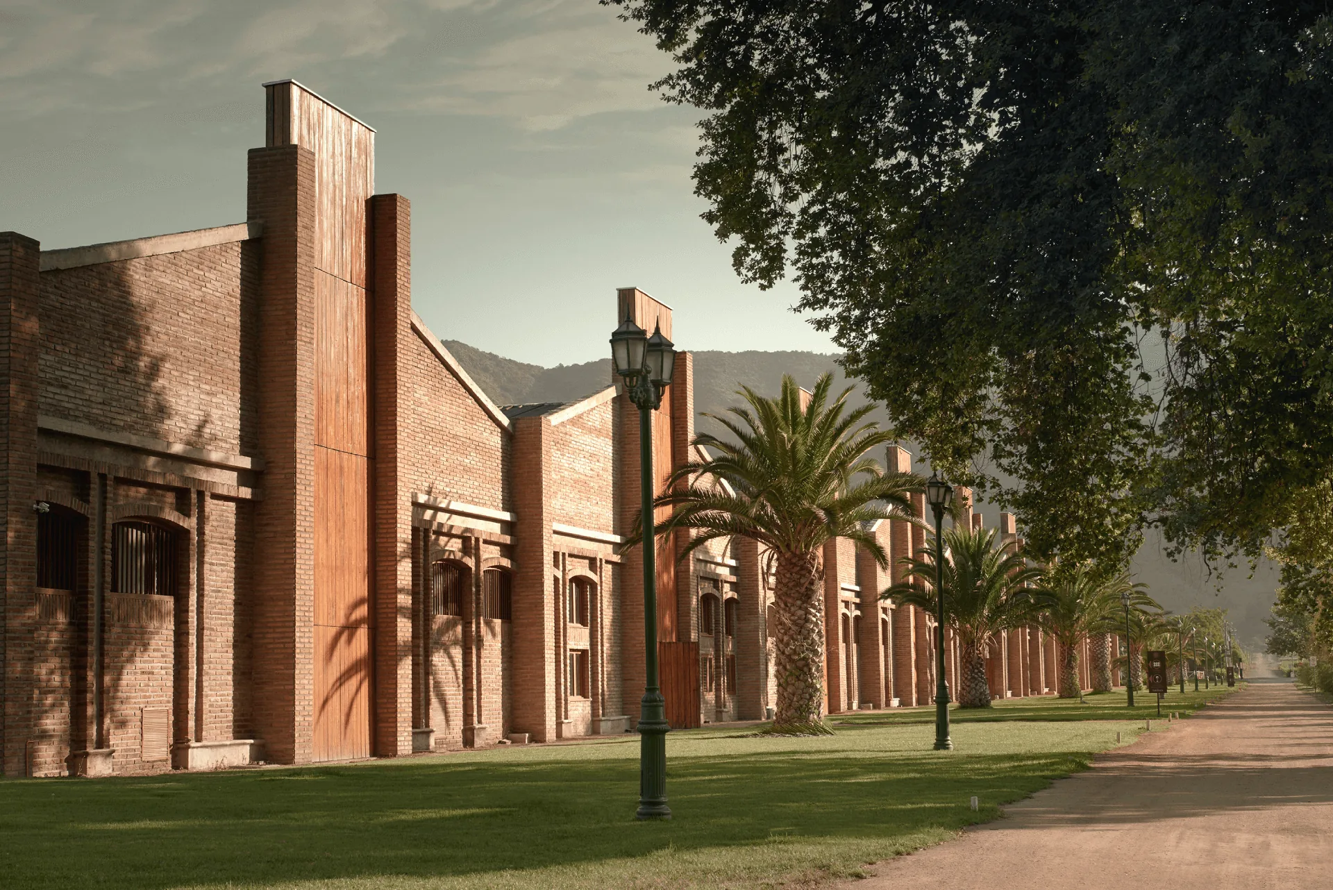 A long red-brick building with tall timber-clad towers beside a lawn and gravel drive, lined with palms and lamp posts