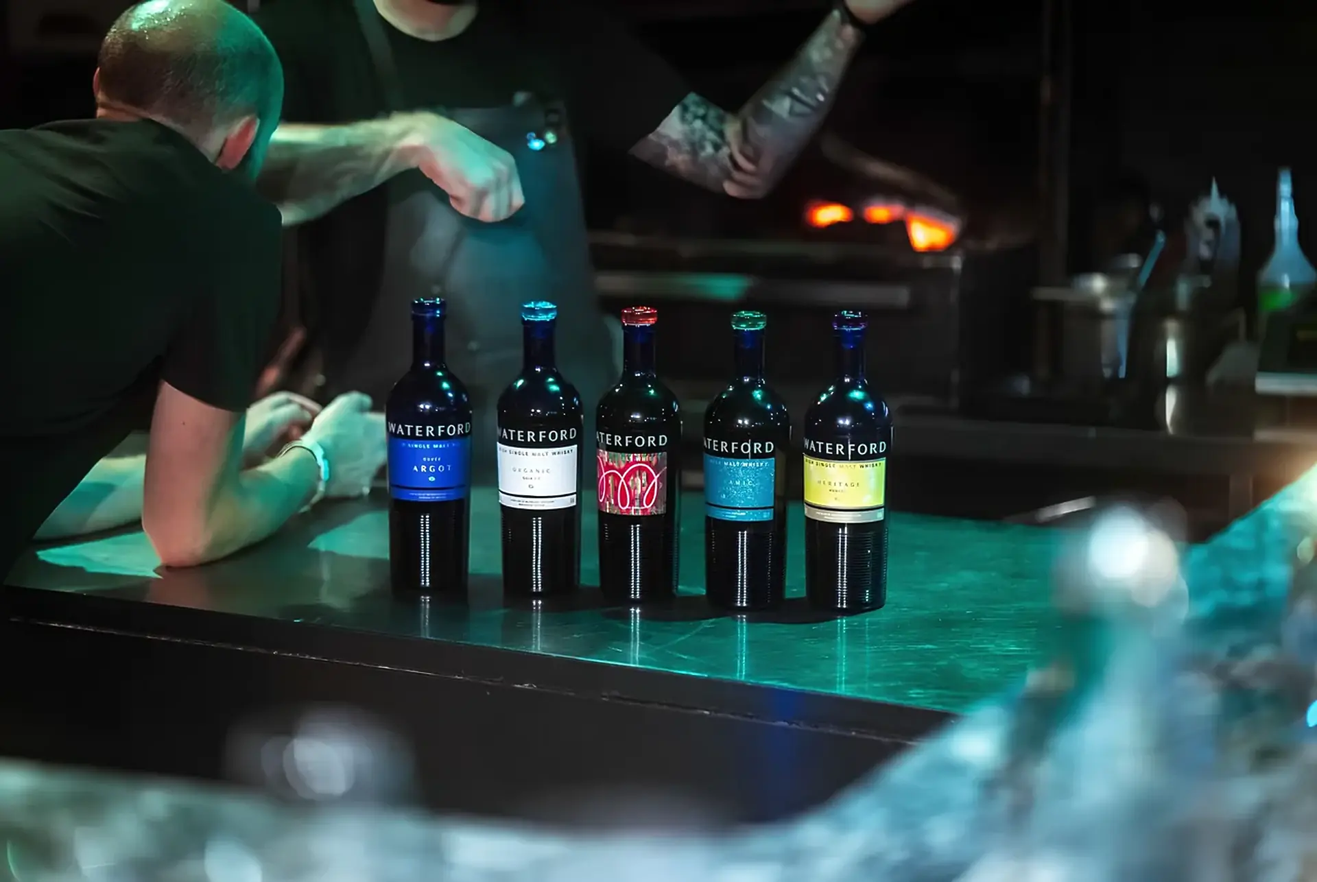 Five Waterford single malt bottles with blue, white, red, teal and yellow labels on a steel counter, two people leaning in, a fire glowing behind