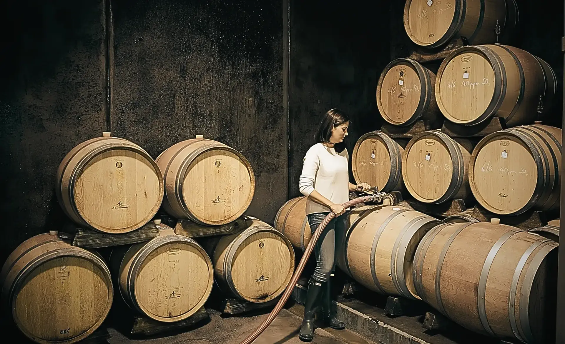 Woman in a white jumper filling an oak barrel through a hose among stacked, chalk-marked barrels in the cellar at A. Christmann