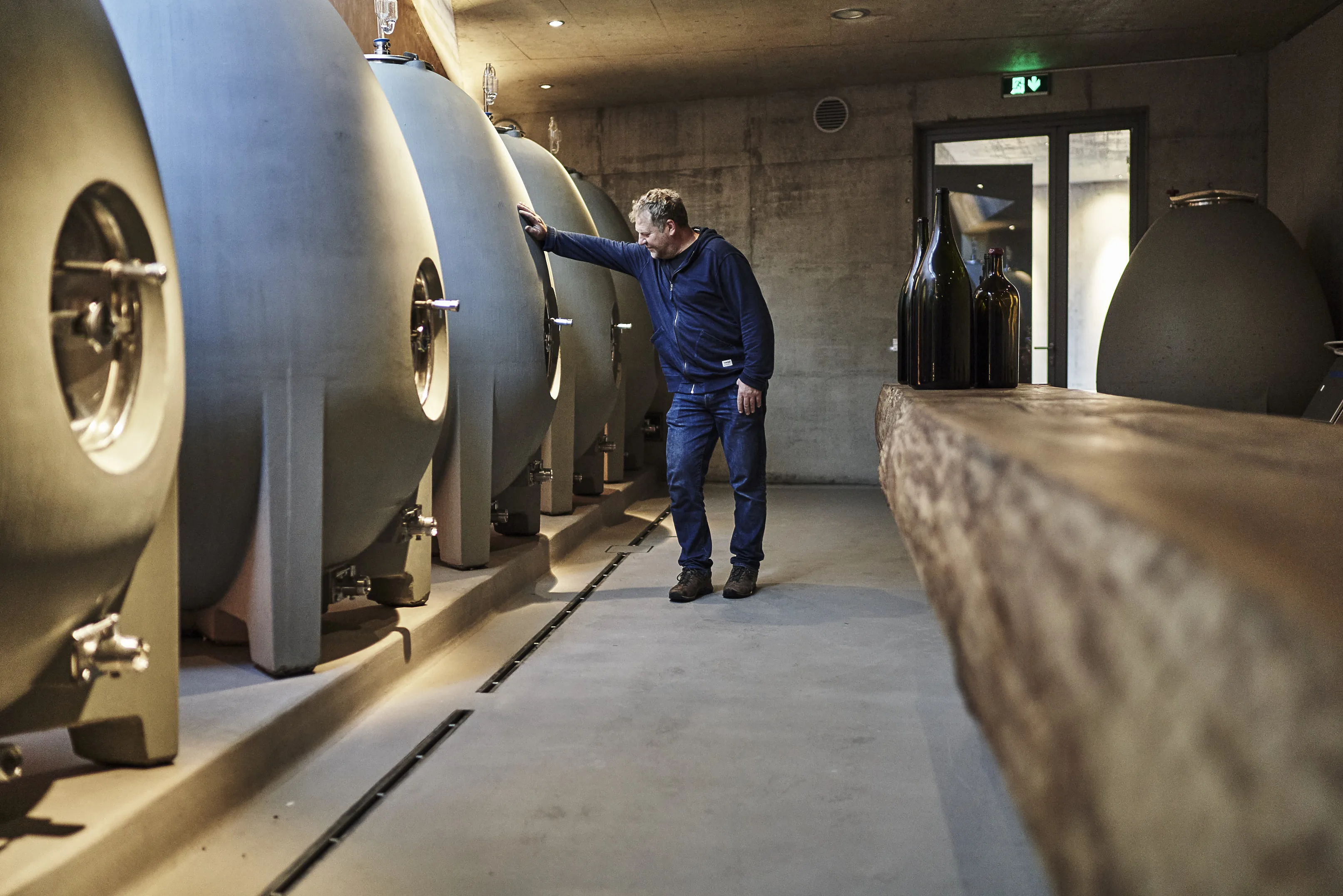 A man resting a hand on a concrete tank in the cellar at Weingut am Stein, with large bottles on a long wooden table