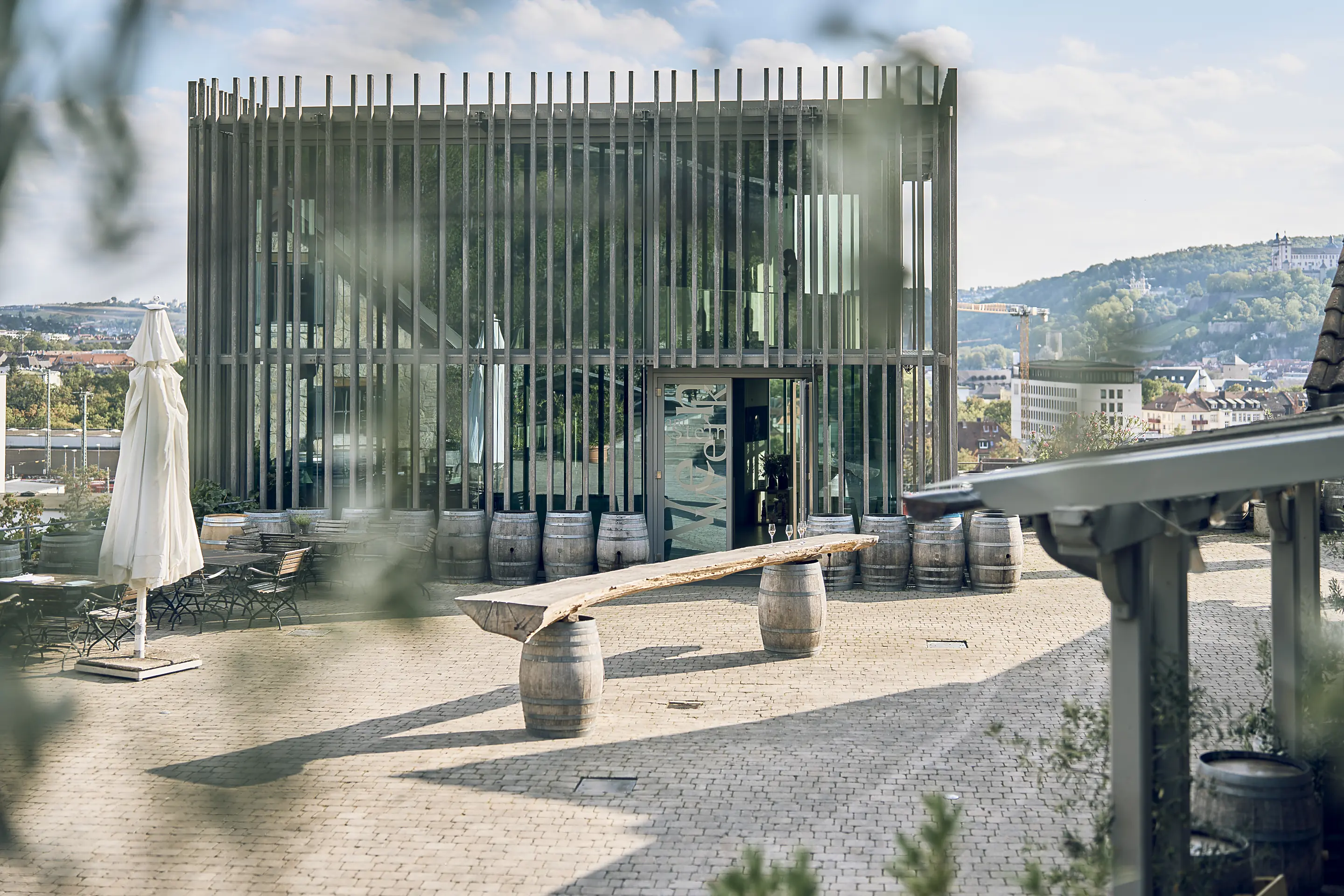 Slatted glass pavilion on the rooftop terrace at Weingut am Stein, with barrels, a long timber bench and Würzburg beyond