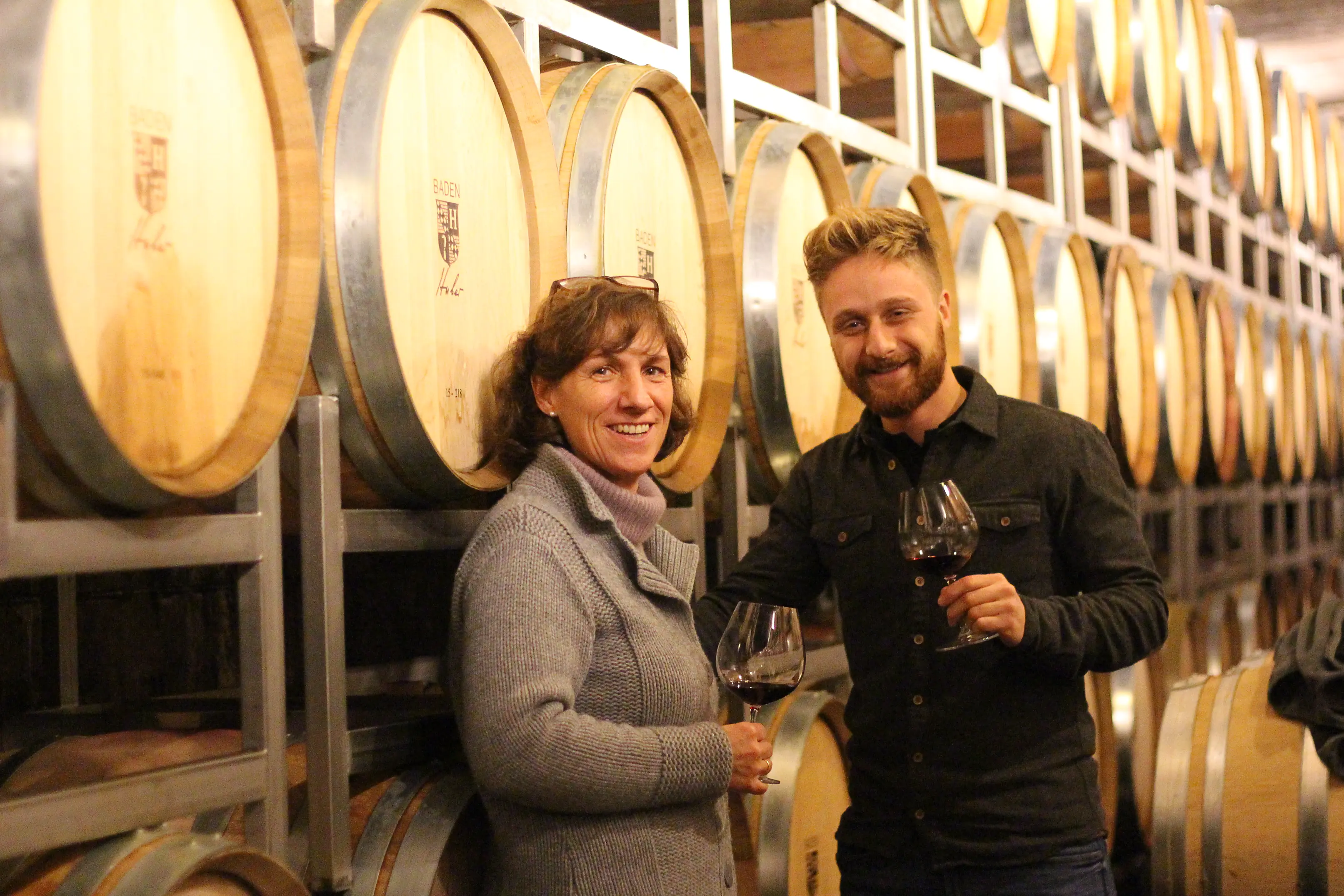 A woman and a bearded young man holding glasses of red wine among stacked barriques branded Huber at Weingut Bernhard Huber
