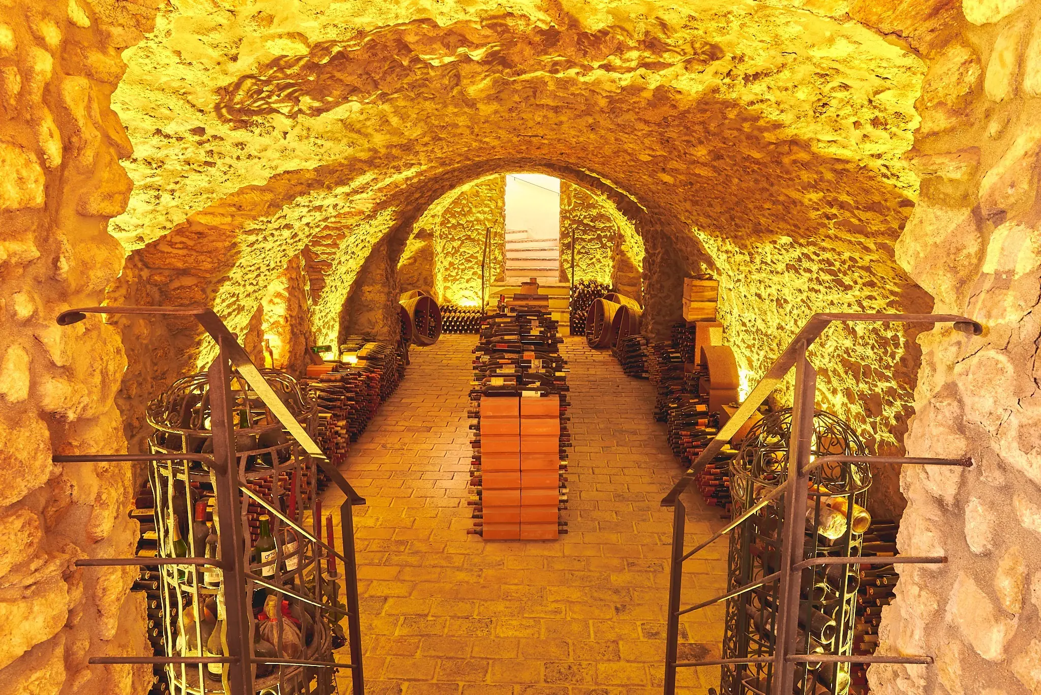 Vaulted stone cellar at Weingut Birgit Braunstein, lit amber, with stacked bottles, iron racks and a stair at the far end