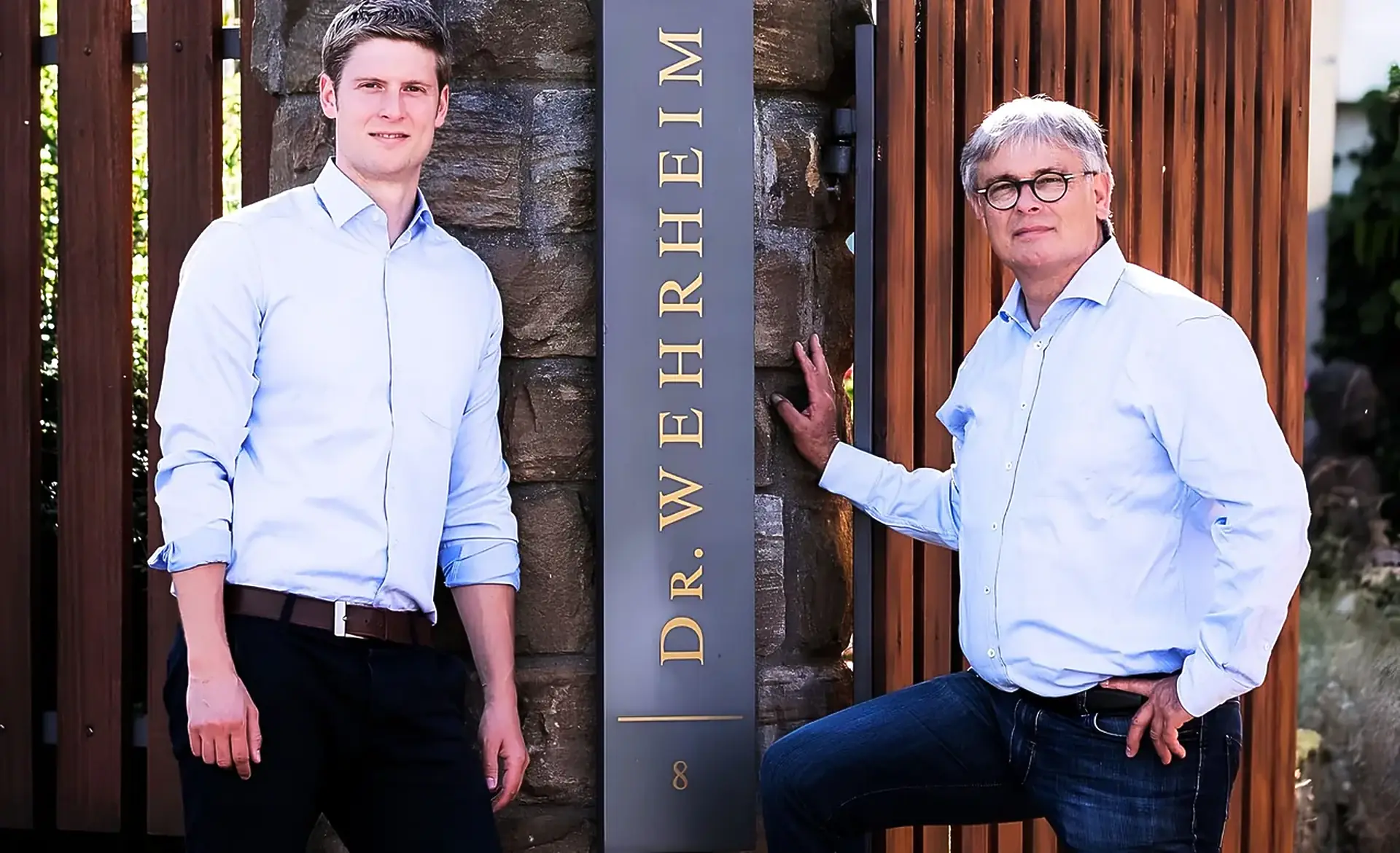 Two men in light blue shirts beside a stone gatepost and a slim sign reading Dr. Wehrheim, with timber slat fencing behind
