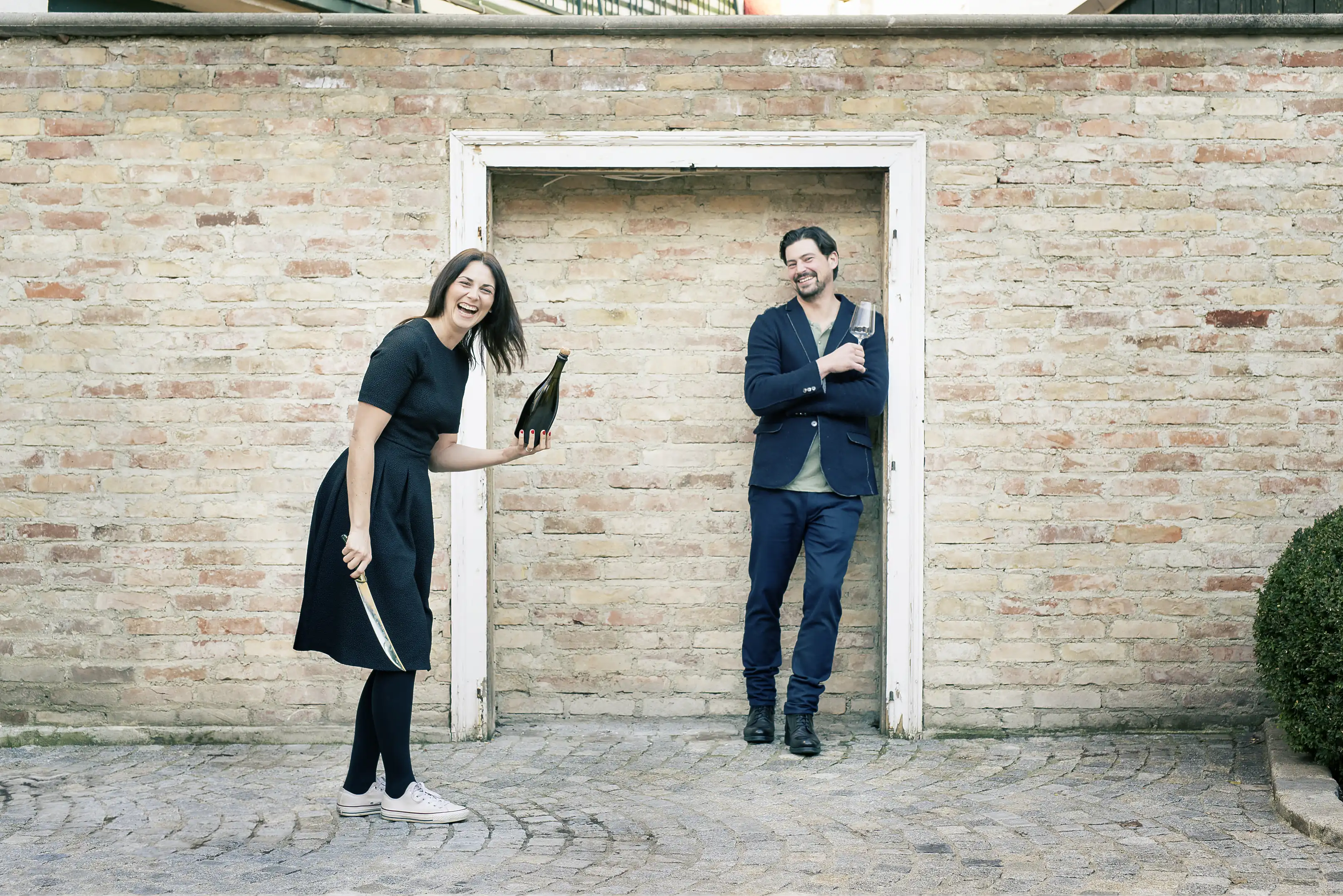 A laughing woman holding a bottle and a sabre and a smiling man holding a wine glass before a brick wall at Weingut Ebner-Ebenauer