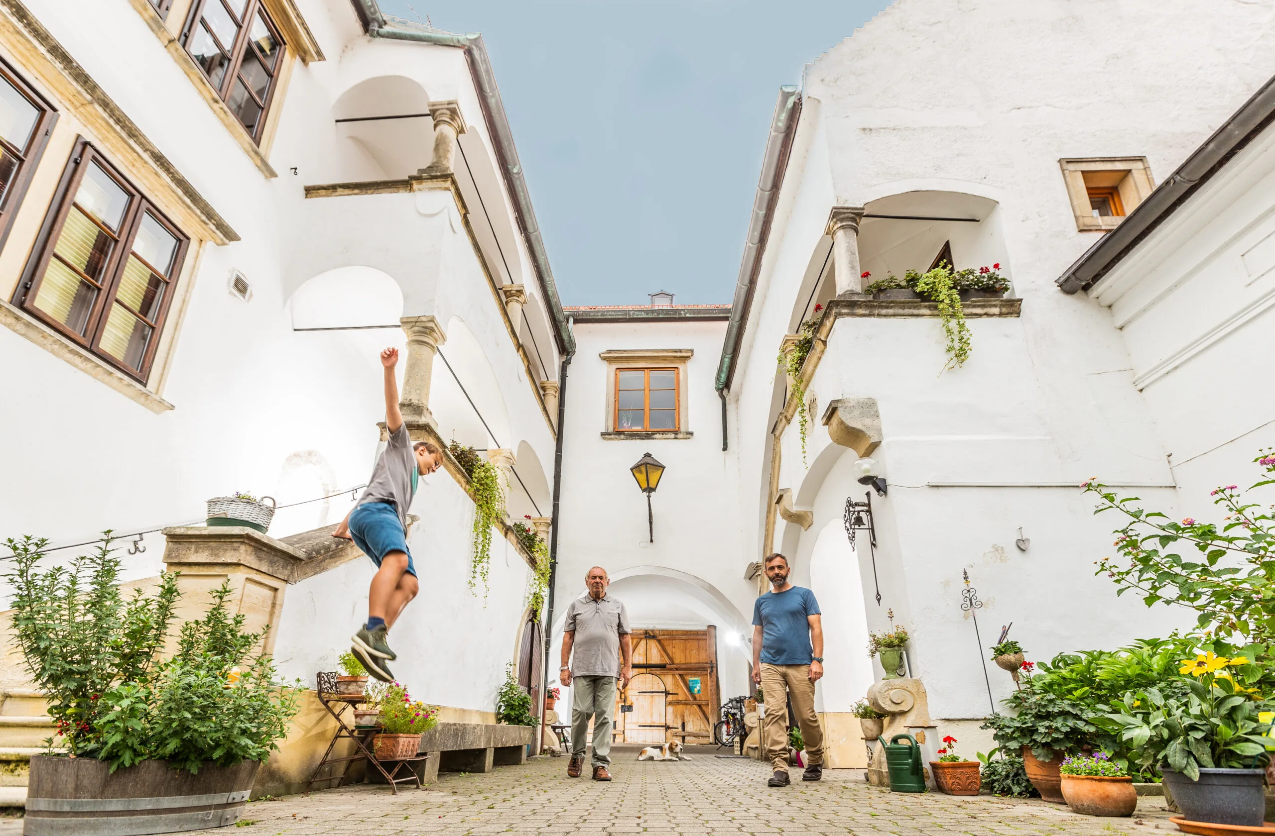 A boy mid-jump in the whitewashed arcaded courtyard at Weingut Feiler-Artinger in Rust, with two men and potted plants
