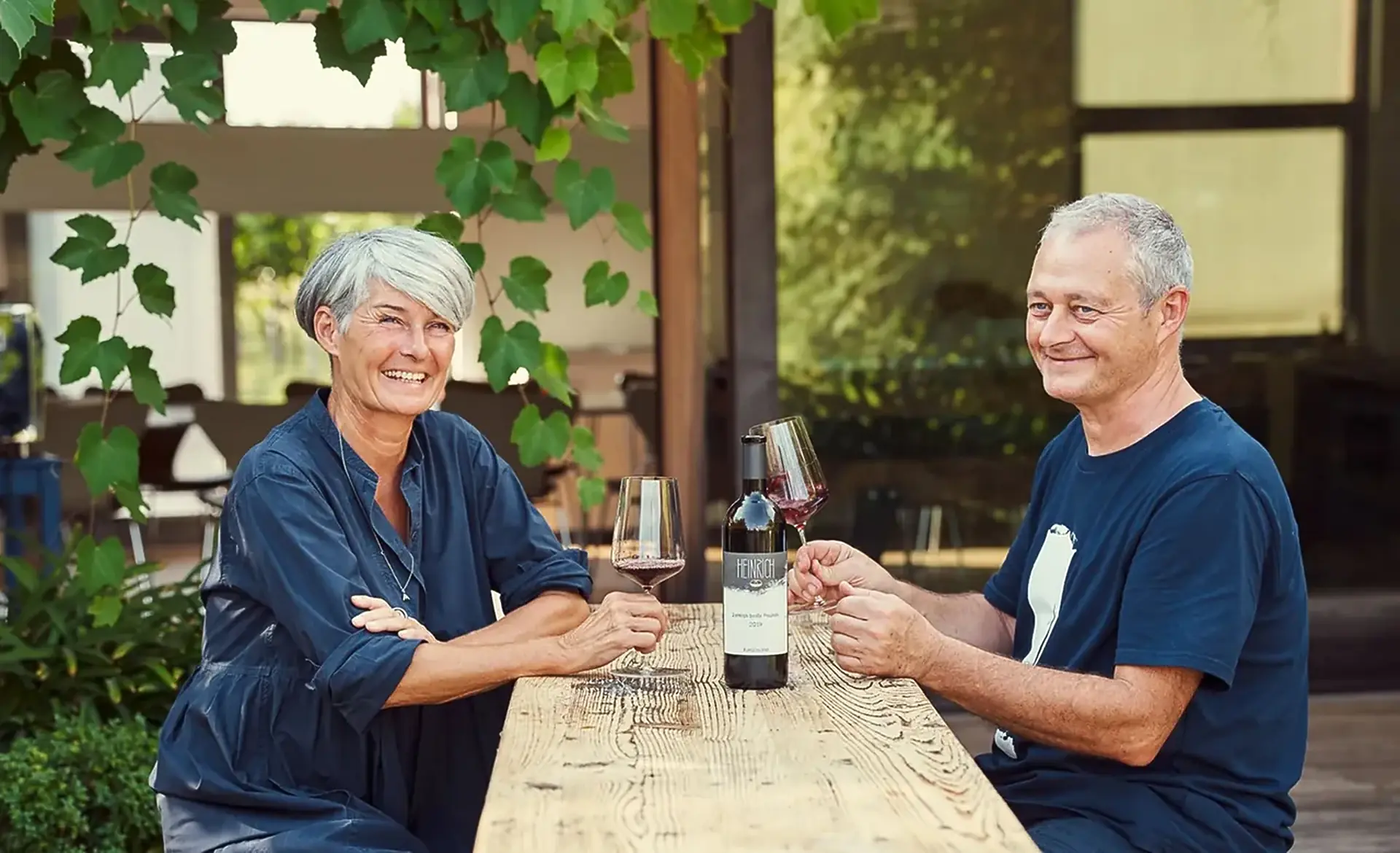 A smiling woman and man at a wooden table under vine leaves, with glasses of red wine and a bottle labelled Heinrich