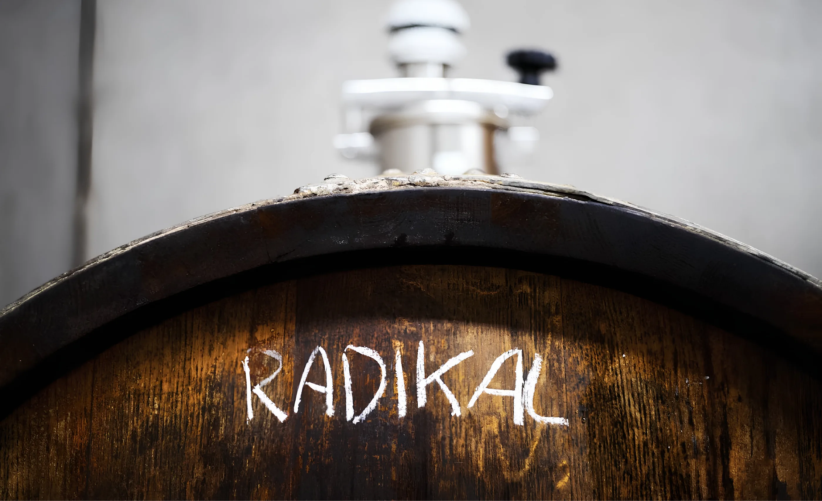 Close view of a dark oak barrel end with RADIKAL chalked on it, a steel valve fitting blurred behind