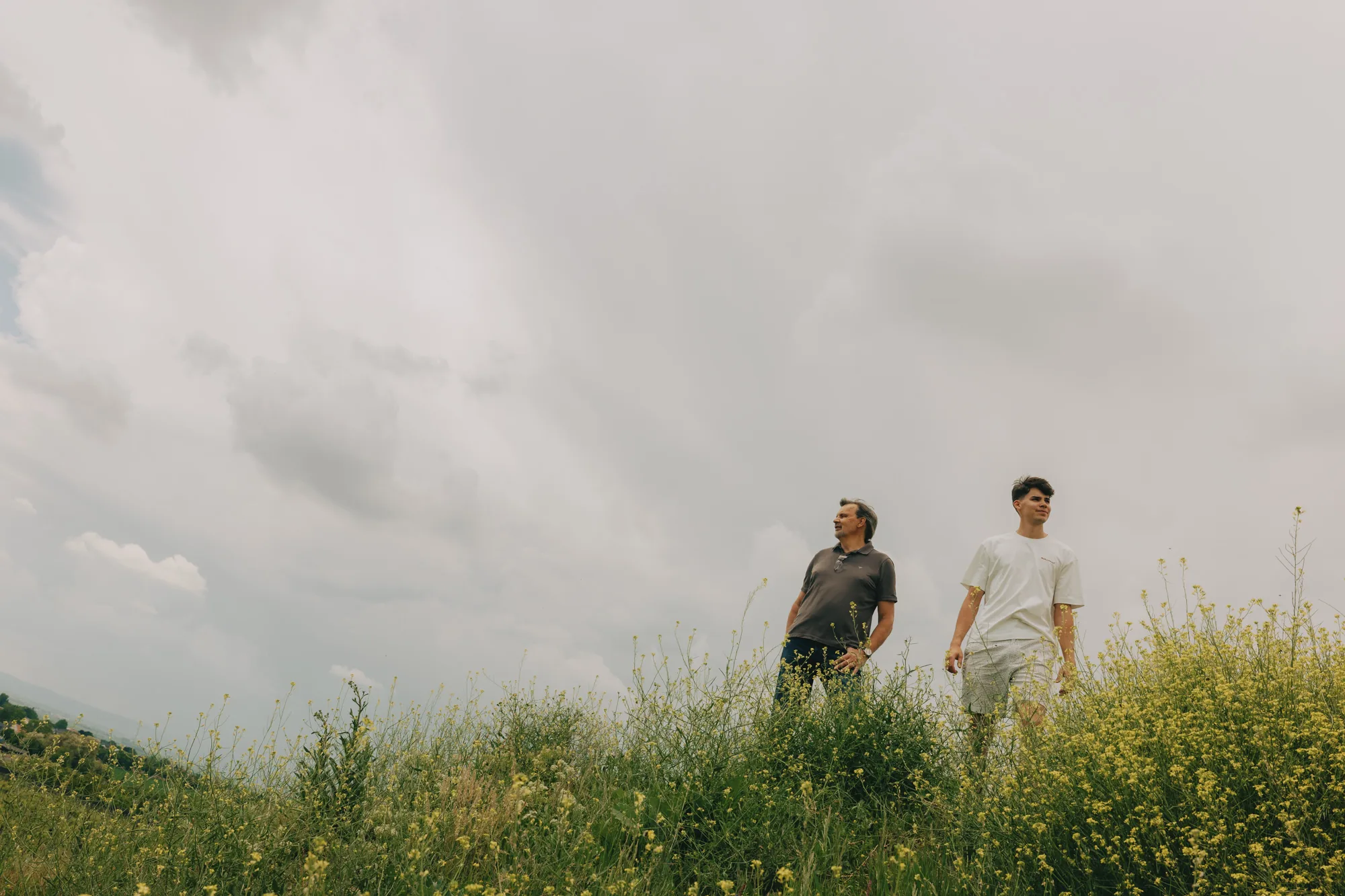 Two men, one older and one younger, standing in tall yellow-flowering weeds on a grassy rise under a heavy cloudy sky