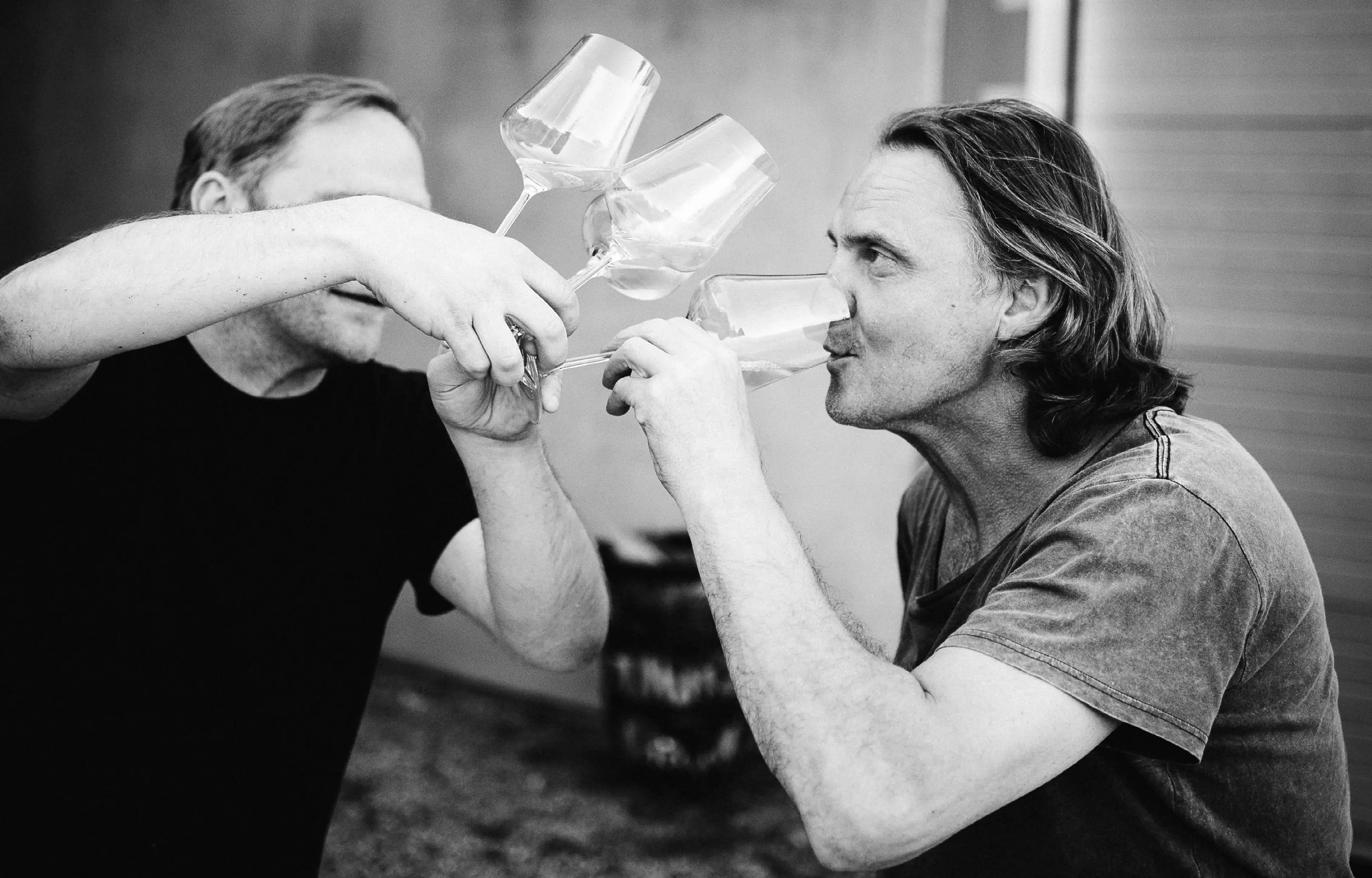 Two men clinking stemmed glasses, one drinking from his glass, in black and white