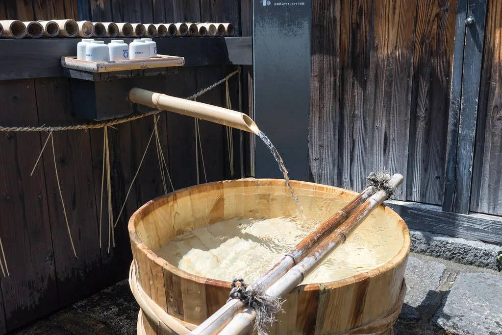 Spring water pouring from a bamboo spout into a cedar basin outside a Japanese brewery